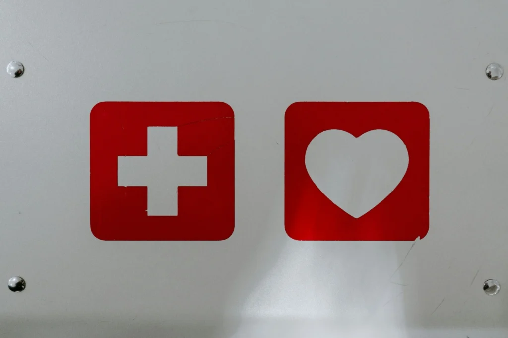 Foto von Blechschild mit zwei Symbolen, einem weißen Kreuzen auf rotem Hintergrund und einem weißen Herz auf rotem Hintergrund. Als Symbol steht das Bild dafür, dass erste Versorgungsangebote endlich eine Art Erste Hilfe für Menschen mit ME/CFS versprechen.