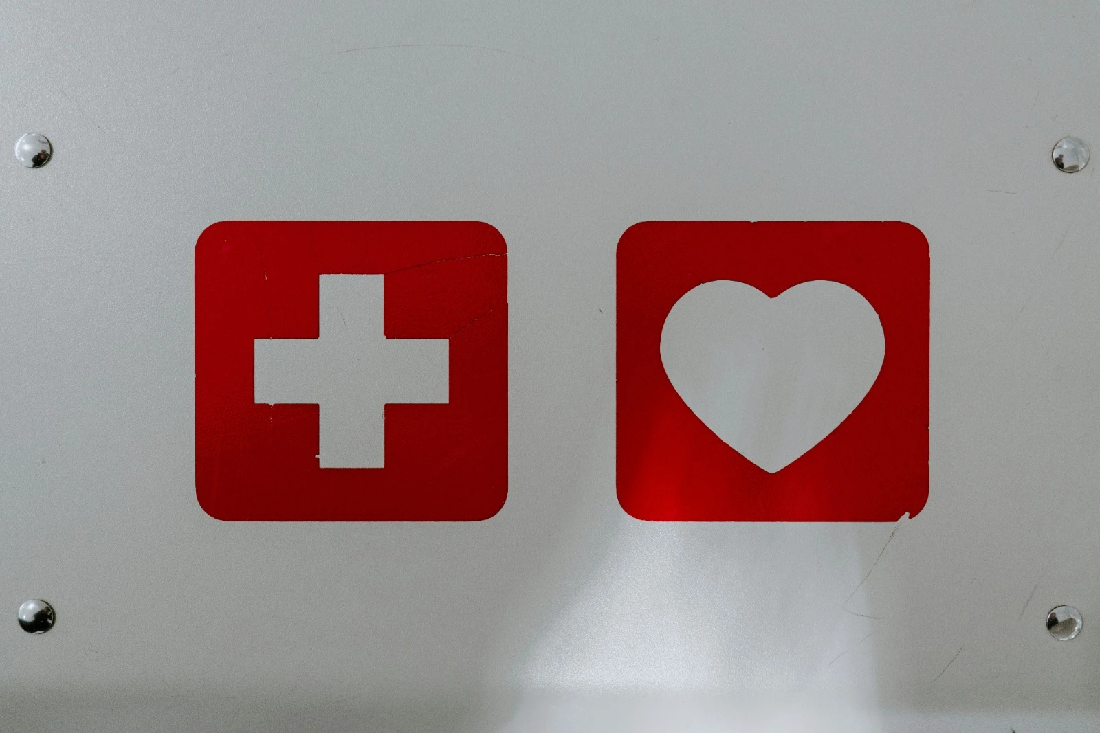Foto von Blechschild mit zwei Symbolen, einem weißen Kreuzen auf rotem Hintergrund und einem weißen Herz auf rotem Hintergrund. Als Symbol steht das Bild dafür, dass erste Versorgungsangebote endlich eine Art Erste Hilfe für Menschen mit ME/CFS versprechen.